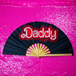 A Bamboo & Silk handheld folding fan that is Black with a Red and White Daddy Print is spread open on a pink sequin background.