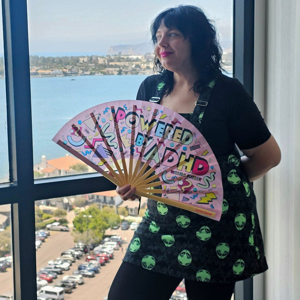 A person with black hair looking out of a hotel window holding a pink silk fan that says Powered by ADHD with a 90's pattern on it