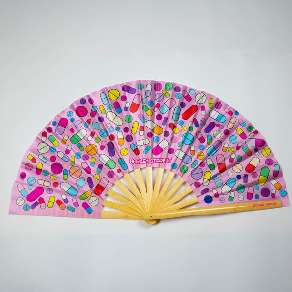 A Bamboo & Silk handheld folding fan that is Pink with a rainbow pill print, is spread open on a white background.