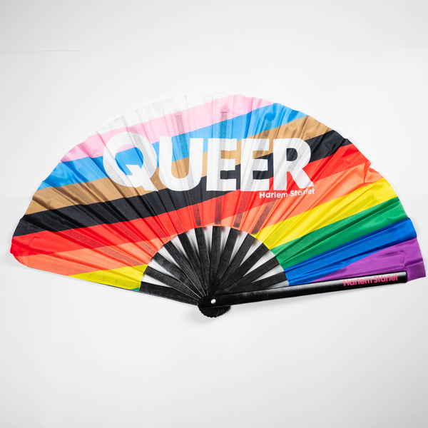 A Bamboo & Silk handheld folding fan with the word Queer written in white over a progress pride flag in diagonal stripes of white, light pink, light blue, brown, black, red, orange, yellow, green, blue and purple is spread open on a white background.