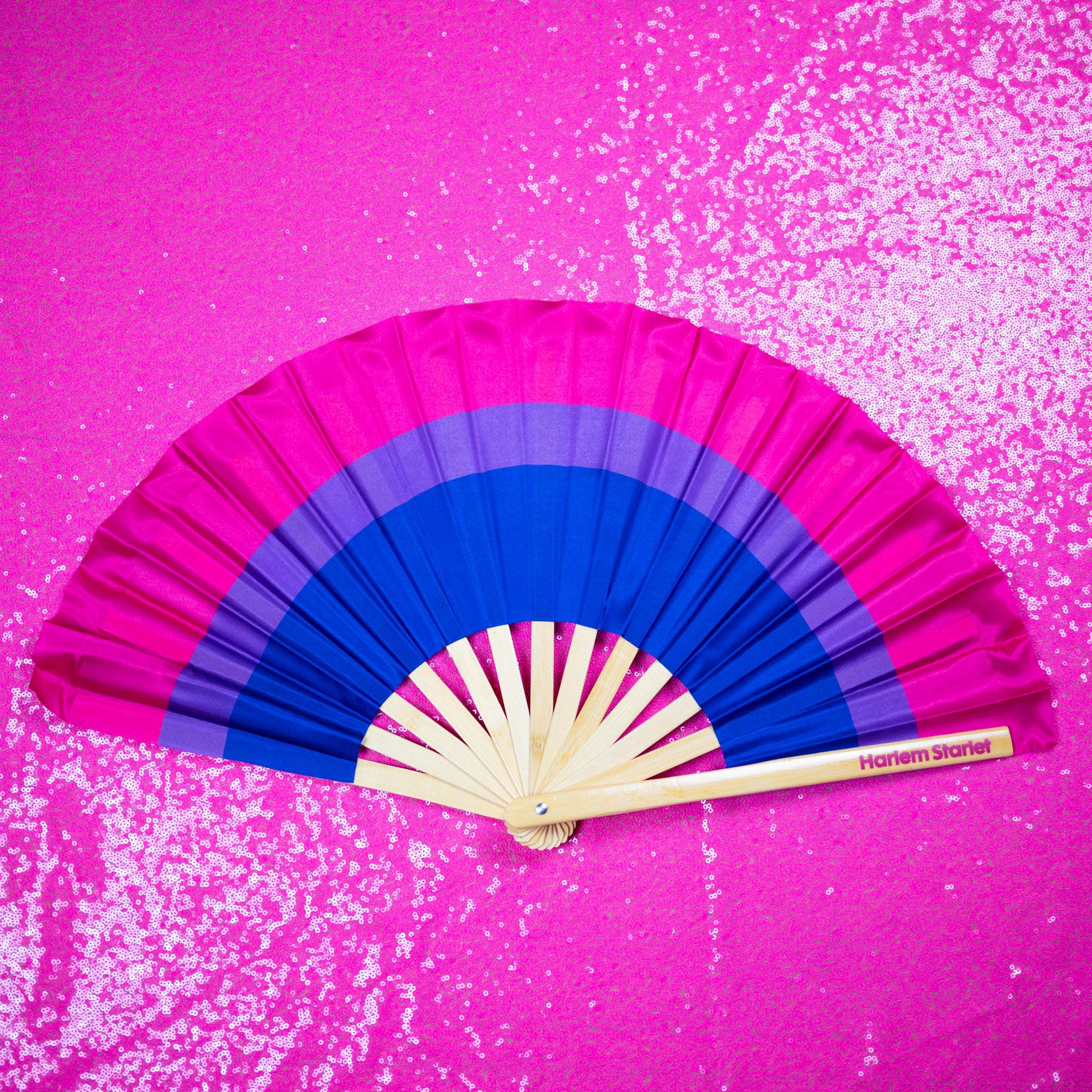 A Bamboo & Silk handheld folding fan that is pink, purple and blue in a bisexual flag design print, is spread open on a pink sequin background.