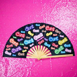 A Bamboo & Silk handheld folding fan that is Black with a rainbow print that says cunt, is spread open on a pink sequin background.