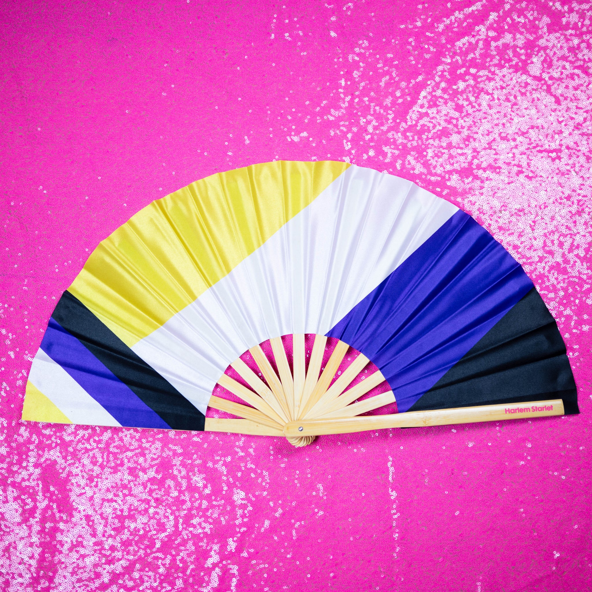 A Bamboo & Silk handheld folding fan that is yellow, white, purple and black in a non binary flag print, is spread open on a pink sequin background.