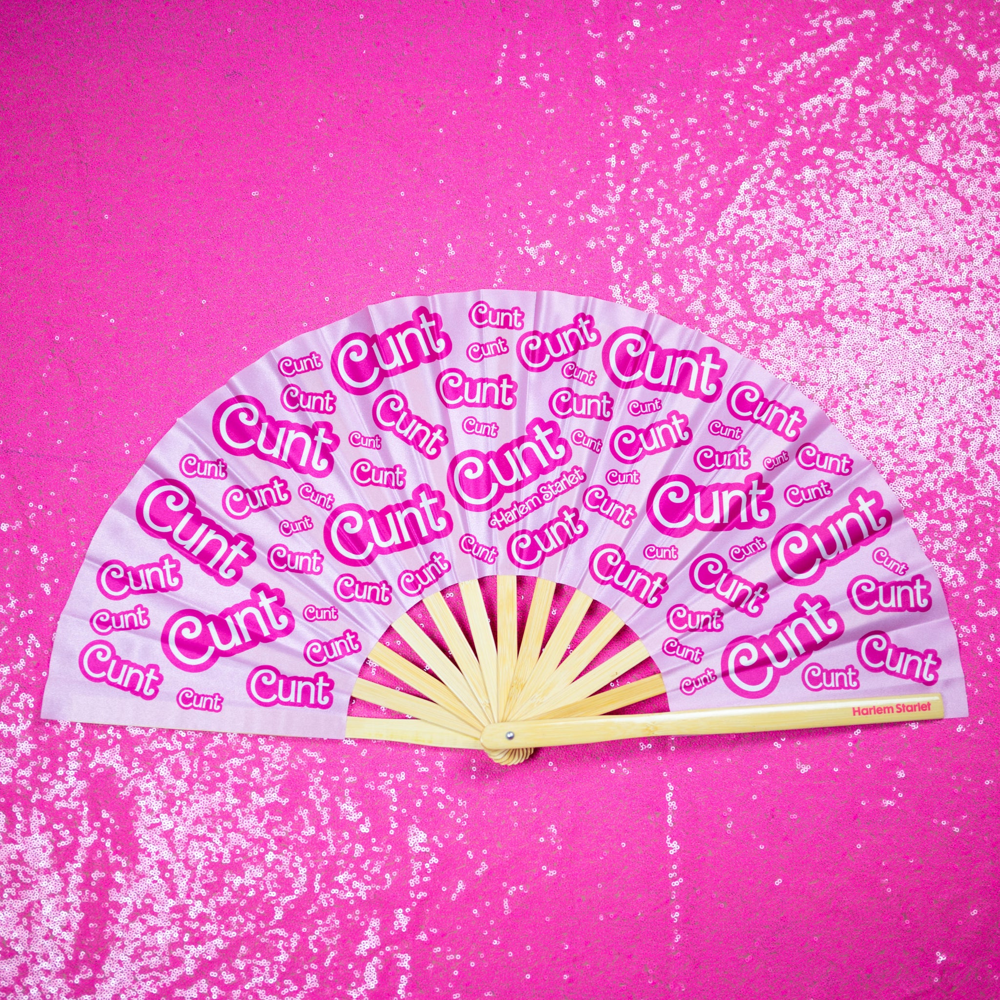 A Bamboo & Silk handheld folding fan that is pink with a hot pink print that says cunt, is spread open on a pink sequin background.