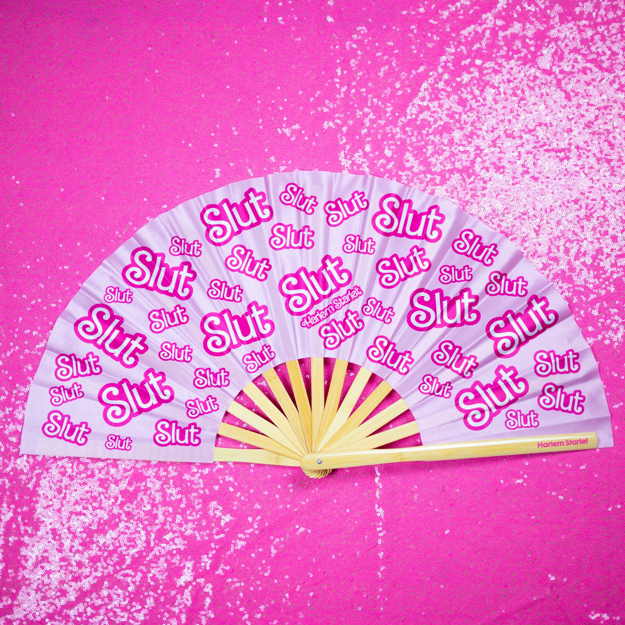 A Bamboo & Silk handheld folding fan that is pink and hot pink, with a repeating slut pattern print, is spread open on a pink sequin background.