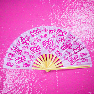 A Bamboo & Silk handheld folding fan that is pink and hot pink, with a repeating slut pattern print, is spread open on a pink sequin background.