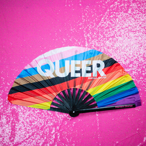 A Bamboo & Silk handheld folding fan with the word Queer written in white over a progress pride flag in diagonal stripes of white, light pink, light blue, brown, black, red, orange, yellow, green, blue and purple is spread open on a pink sequin background.