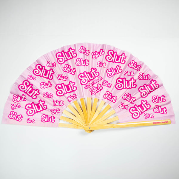 A Bamboo & Silk handheld folding fan that is pink and hot pink, with a repeating slut pattern print, is spread open on a pink sequin background.