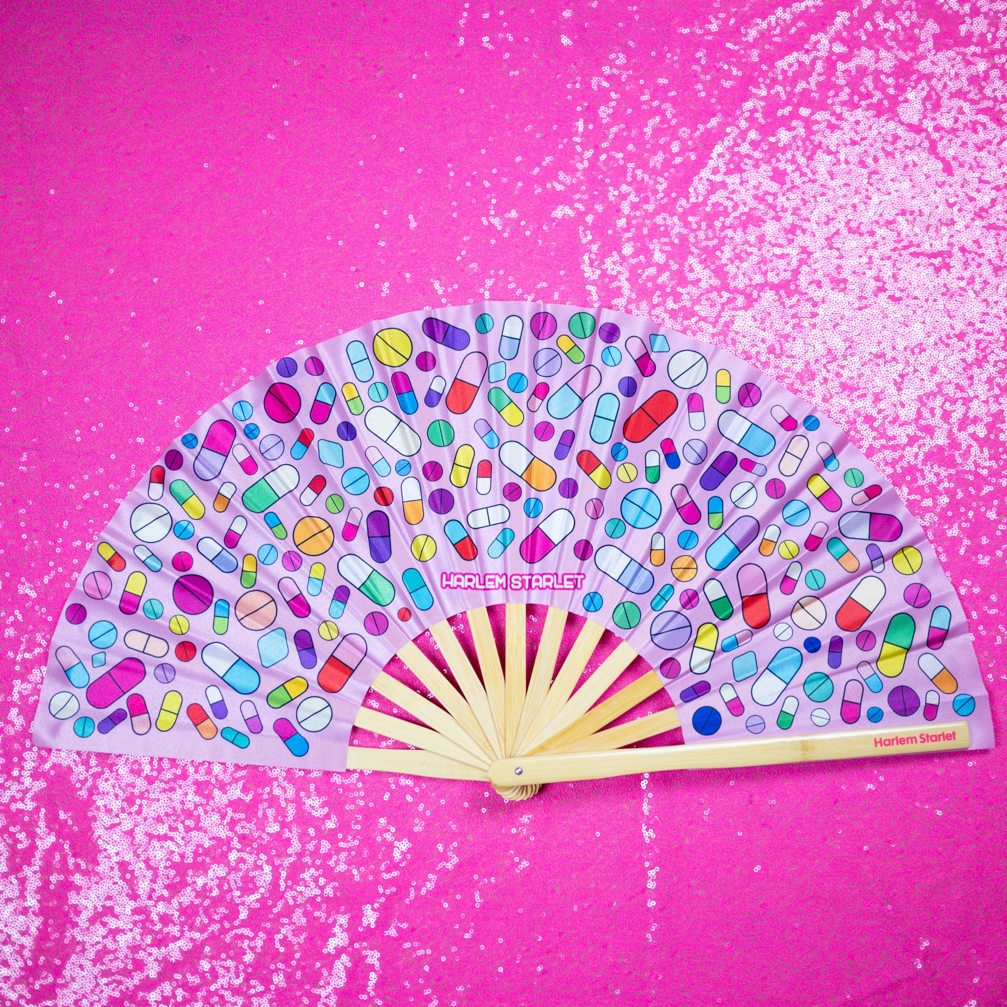 A Bamboo & Silk handheld folding fan that is Pink with a rainbow pill print, is spread open on a pink sequin background.