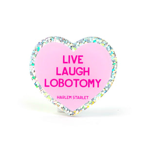 Heart pin with a pink background, featuring hot pink text that says "Live, Laugh, Lobotomy". Edges are silver with multicolored glitter. Mental Health Pins.