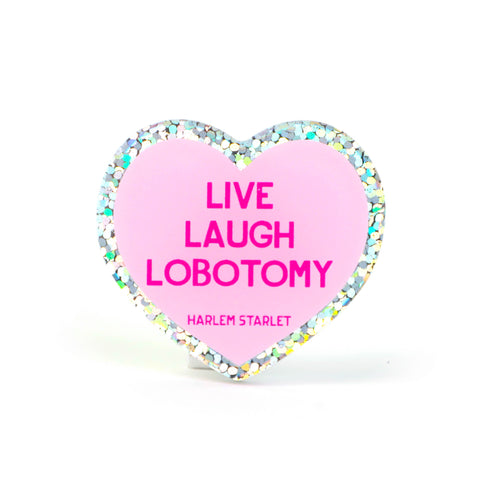 Heart pin with a pink background, featuring hot pink text that says "Live, Laugh, Lobotomy". Edges are silver with multicolored glitter. Mental Health Pins.