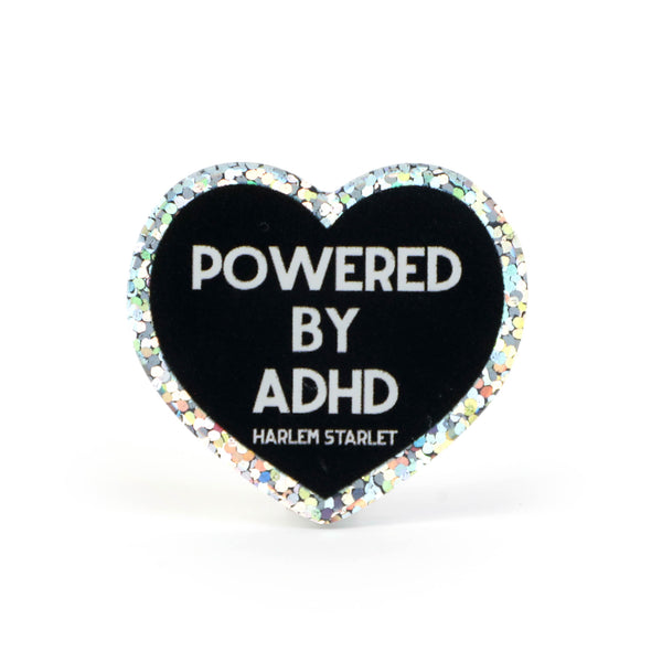 Heart pin with a black background, featuring white text that says "Powered by ADHD!". Edges are silver with multicolored glitter. Neurodivergent Pride Pin.