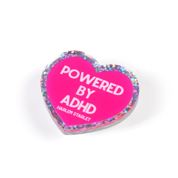 Heart pin with a hot pink background, featuring light pink text that says "Powered by ADHD!". Edges are silver with multicolored glitter. Neurodivergent Pride Pin.