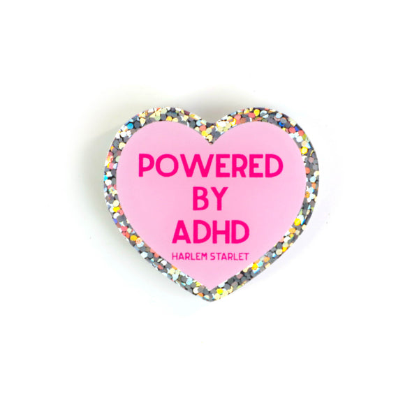 Heart pin with a pink background, featuring hot pink text that says "Powered by ADHD!". Edges are silver with multicolored glitter. Neurodivergent Pride Pin.