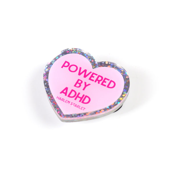 Heart pin with a pink background, featuring hot pink text that says "Powered by ADHD!". Edges are silver with multicolored glitter. Neurodivergent Pride Pin.