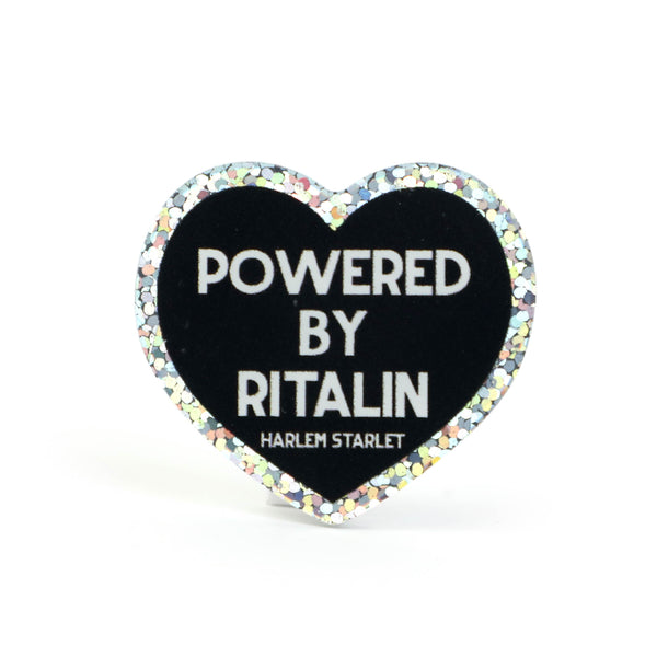 Heart pin with black background, featuring white text that says "Powered by Ritalin". Edges are silver with holographic glitter. Neurodivergent Pride Pin.