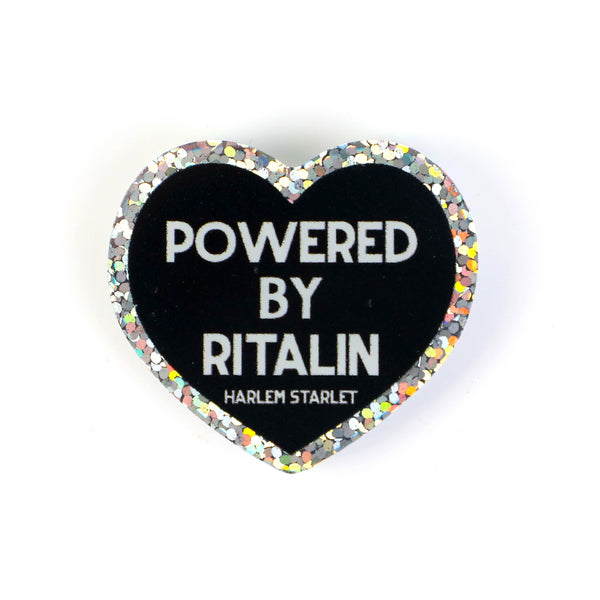 Heart pin with black background, featuring white text that says "Powered by Ritalin". Edges are silver with holographic glitter. Neurodivergent Pride Pin.