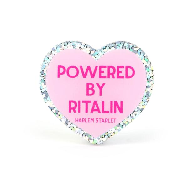 Heart pin with a pink background, featuring hot pink text that says "Powered by Ritalin". Edges are silver with multicolored glitter. Neurodivergent Pride Pin.