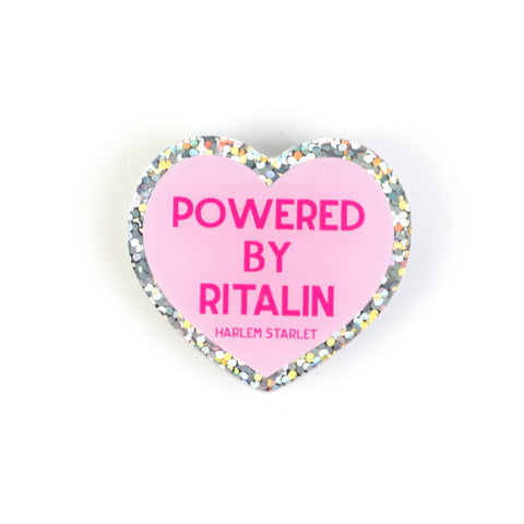 Heart pin with a pink background, featuring hot pink text that says "Powered by Ritalin". Edges are silver with multicolored glitter. Neurodivergent Pride Pin.