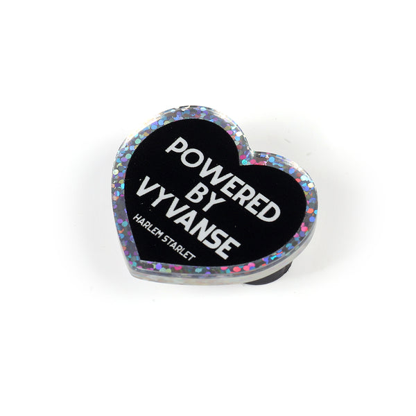 Heart pin with black background, featuring white text that says "Powered by Vyvanse". Edges are silver with holographic glitter. Neurodivergent Pride Pin.