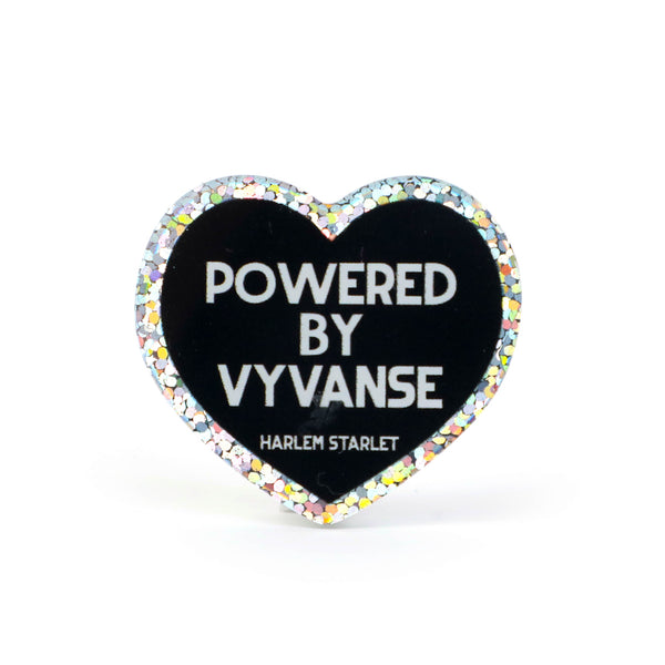 Heart pin with black background, featuring white text that says "Powered by Vyvanse". Edges are silver with holographic glitter. Neurodivergent Pride Pin.