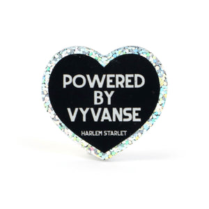 Heart pin with black background, featuring white text that says "Powered by Vyvanse". Edges are silver with holographic glitter. Neurodivergent Pride Pin.