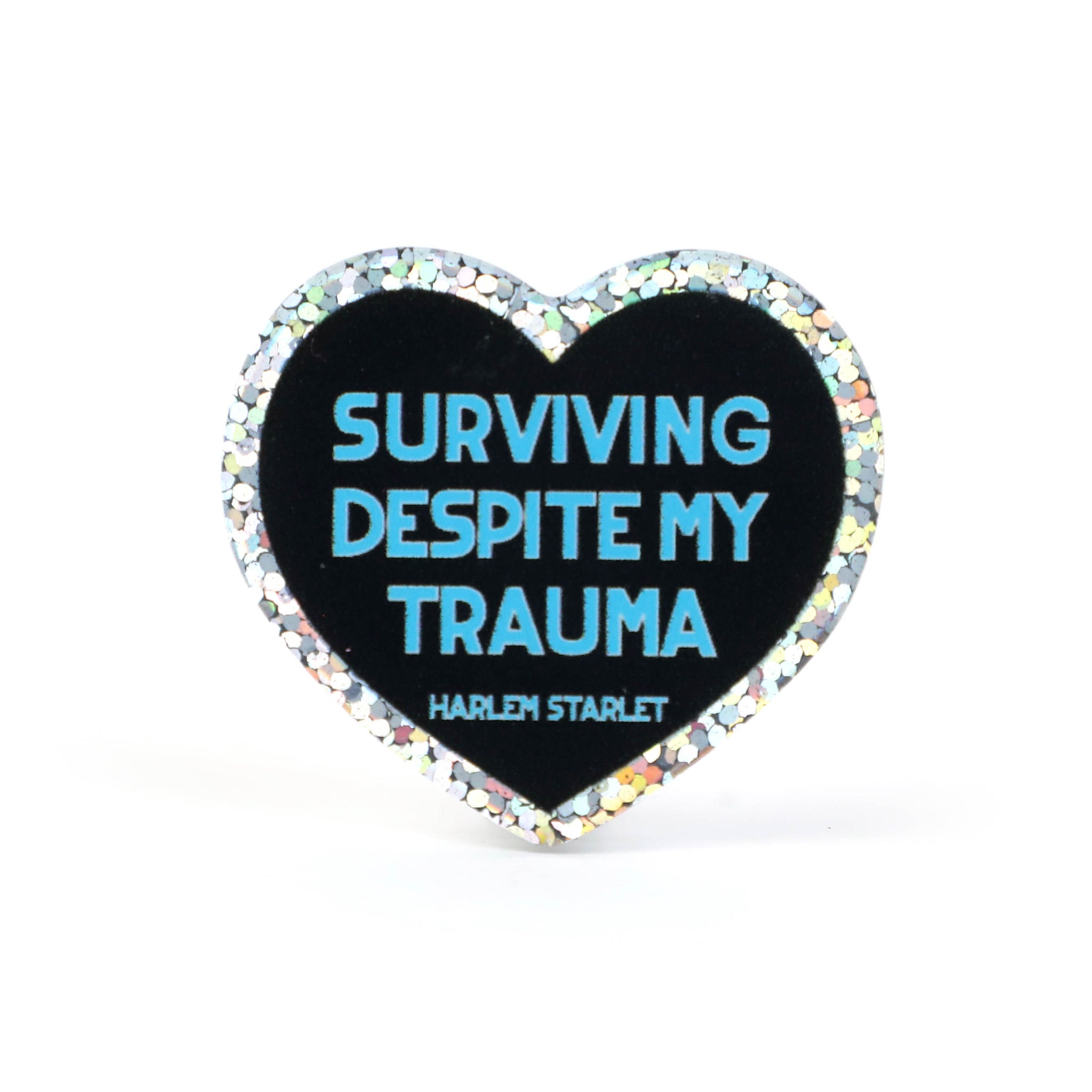 Heart pin with a black background, featuring light blue text that says "Surviving Despite My Trauma". Edges are silver with multicolored glitter.