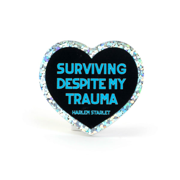 Heart pin with a black background, featuring light blue text that says "Surviving Despite My Trauma". Edges are silver with multicolored glitter.