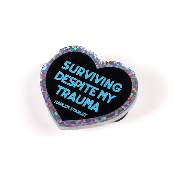 Heart pin with a black background, featuring light blue text that says "Surviving Despite My Trauma". Edges are silver with multicolored glitter.