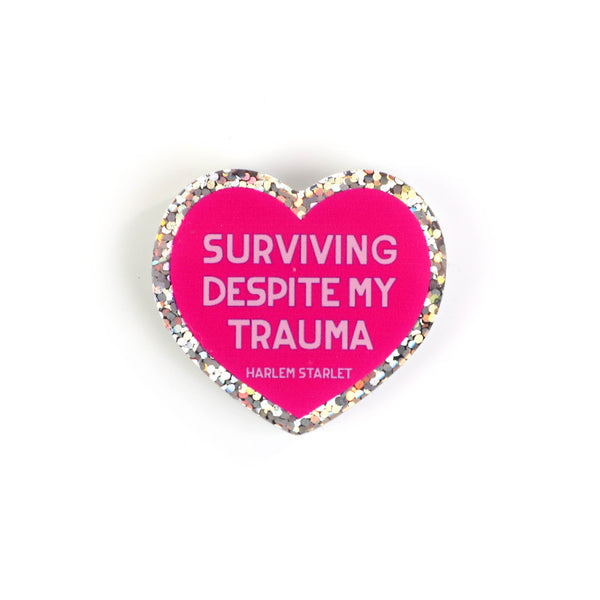 Heart pin with a hot pink background, featuring light pink text that says "Surviving Despite My Trauma". Edges are silver with multicolored glitter.