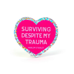 Heart pin with a hot pink background, featuring light pink text that says "Surviving Despite My Trauma". Edges are silver with multicolored glitter.