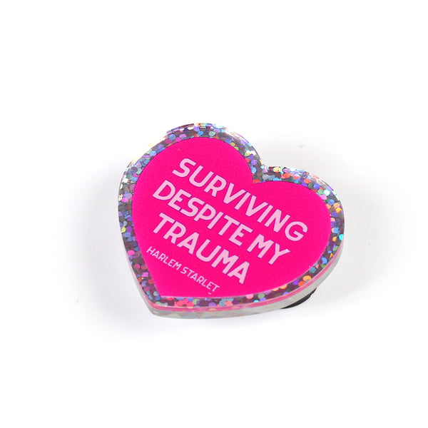 Heart pin with a hot pink background, featuring light pink text that says "Surviving Despite My Trauma". Edges are silver with multicolored glitter.