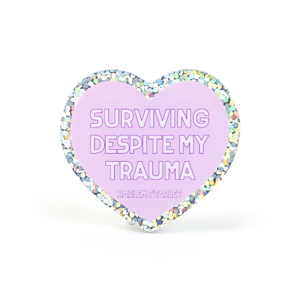 Heart pin with a lilac background, featuring white text that says "Surviving Despite My Trauma". Edges are silver with multicolored glitter.