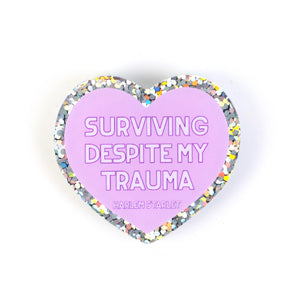 featuring white text that says "Surviving Despite My Trauma". Edges are silver with multicolored glitter.