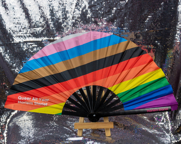 A Bamboo & Silk handheld folding fan with the word Queer All Year written in white over a progress pride flag in diagonal stripes of white, light pink, light blue, brown, black, red, orange, yellow, green, blue and purple is spread open on a silver sequin background.